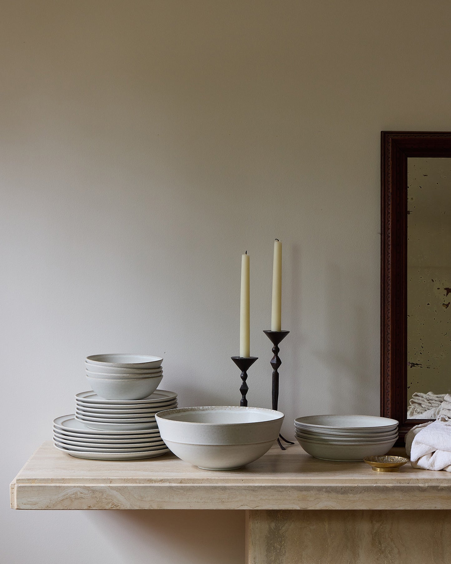 Set of stoneware dishes on a travertine table with candles and a mirror in the background.