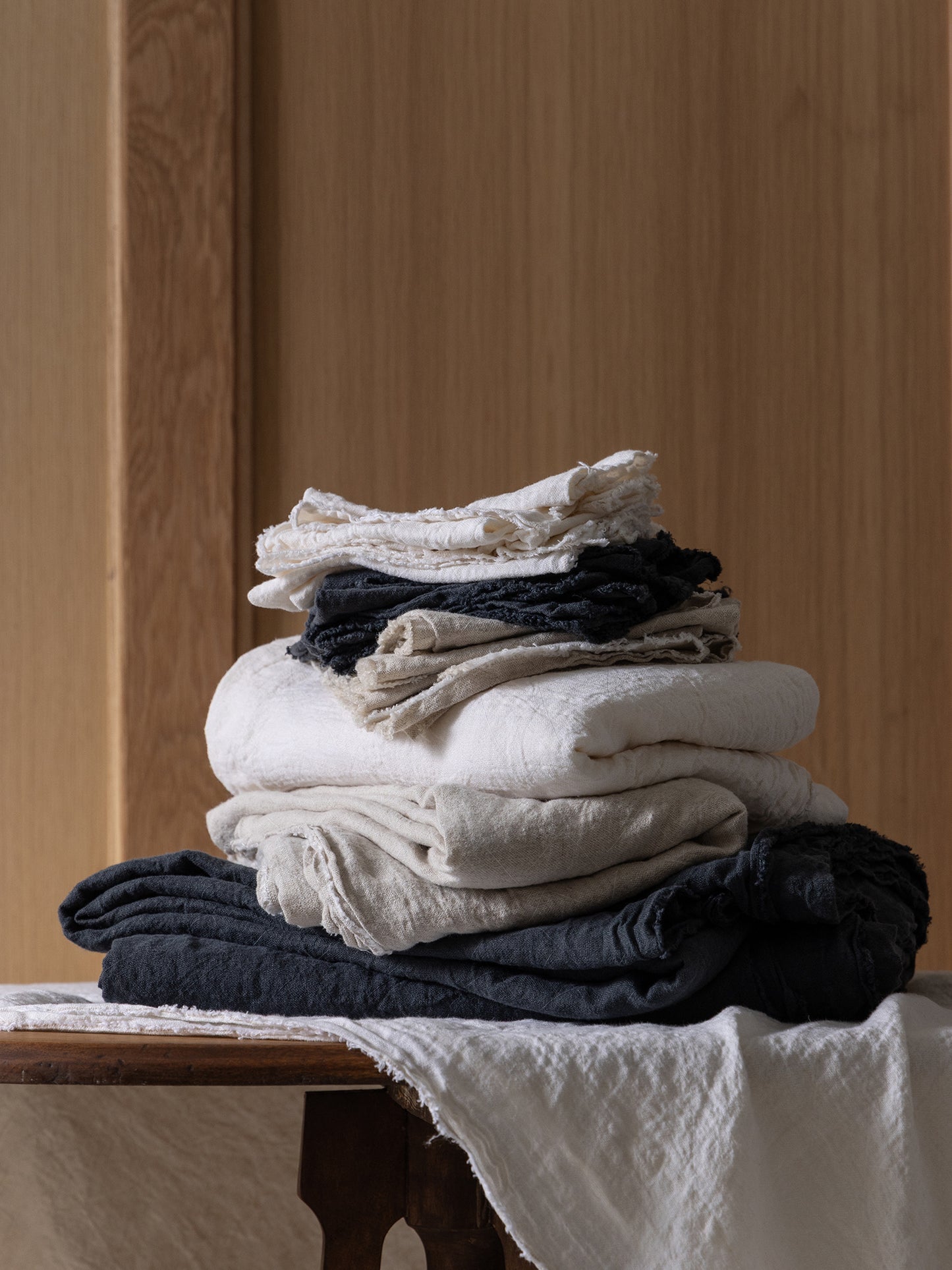 Stack of white, natural, and dark grey linen napkins and tablecloths on wood table against wood wall.