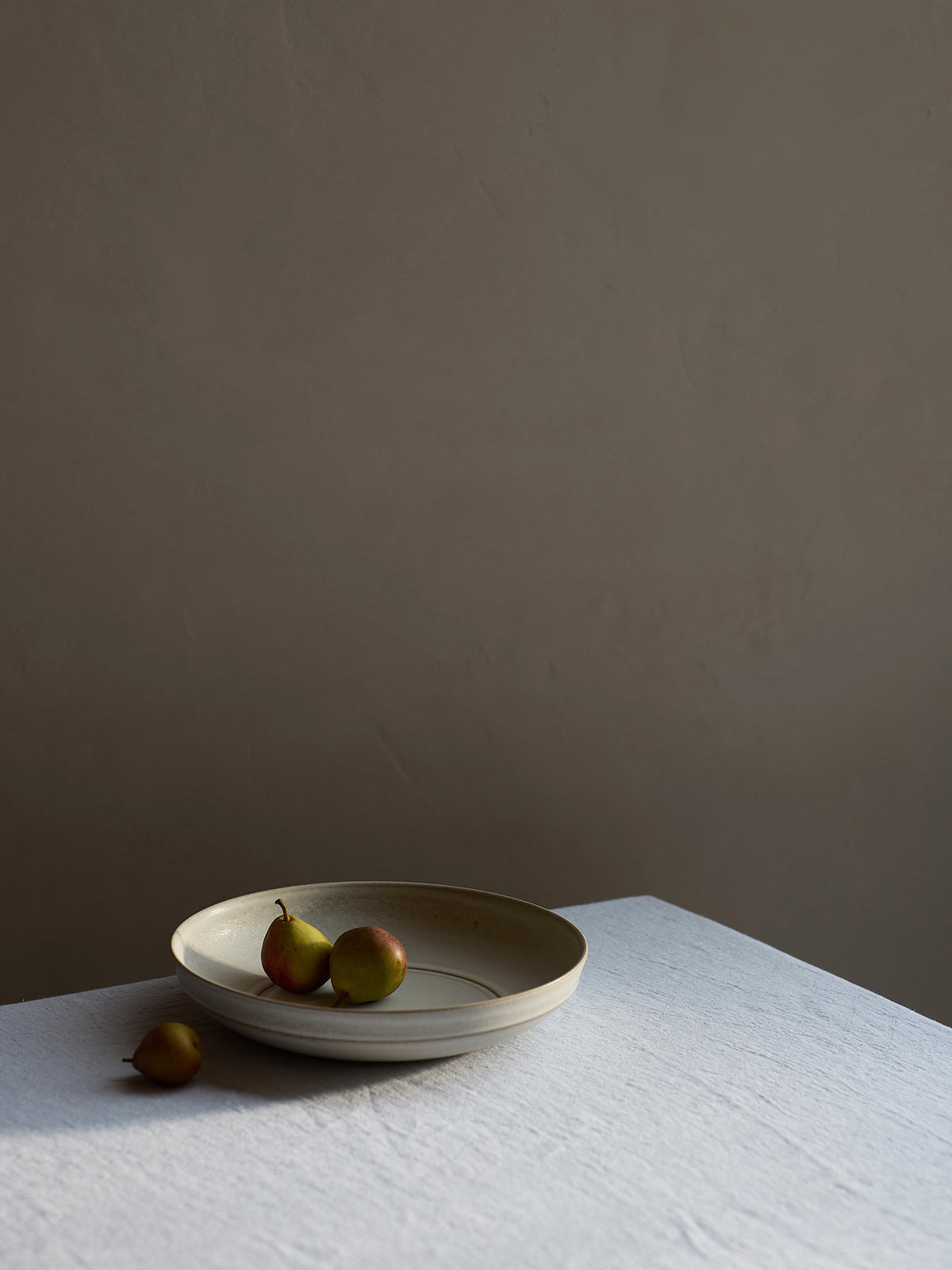 A shallow round ceramic serving bowl in a warm beige tone, holding two small seckel pears, set on a white textured tablecloth against a dark taupe wall.