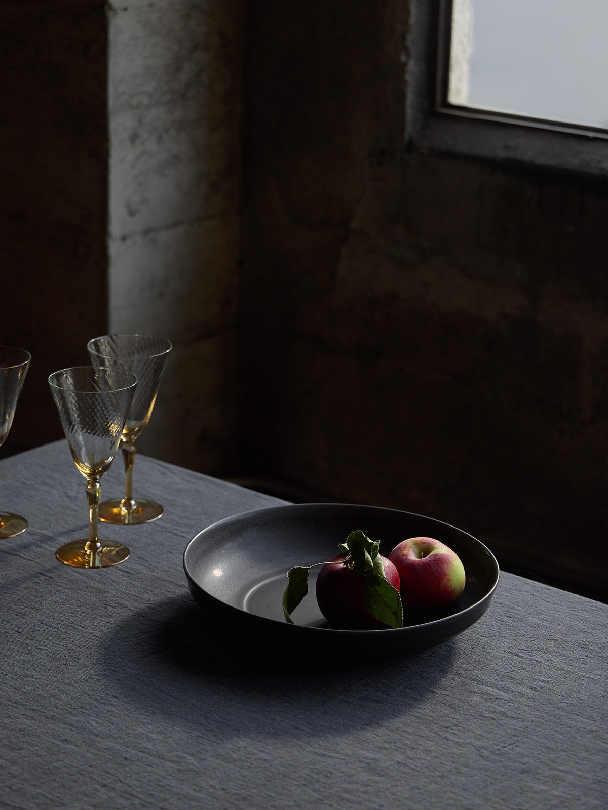 A dark gray ceramic bowl containing two red apples with green leaves, styled on a slate tablecloth next to amber toned wine glasses in a dimly lit industrial room.