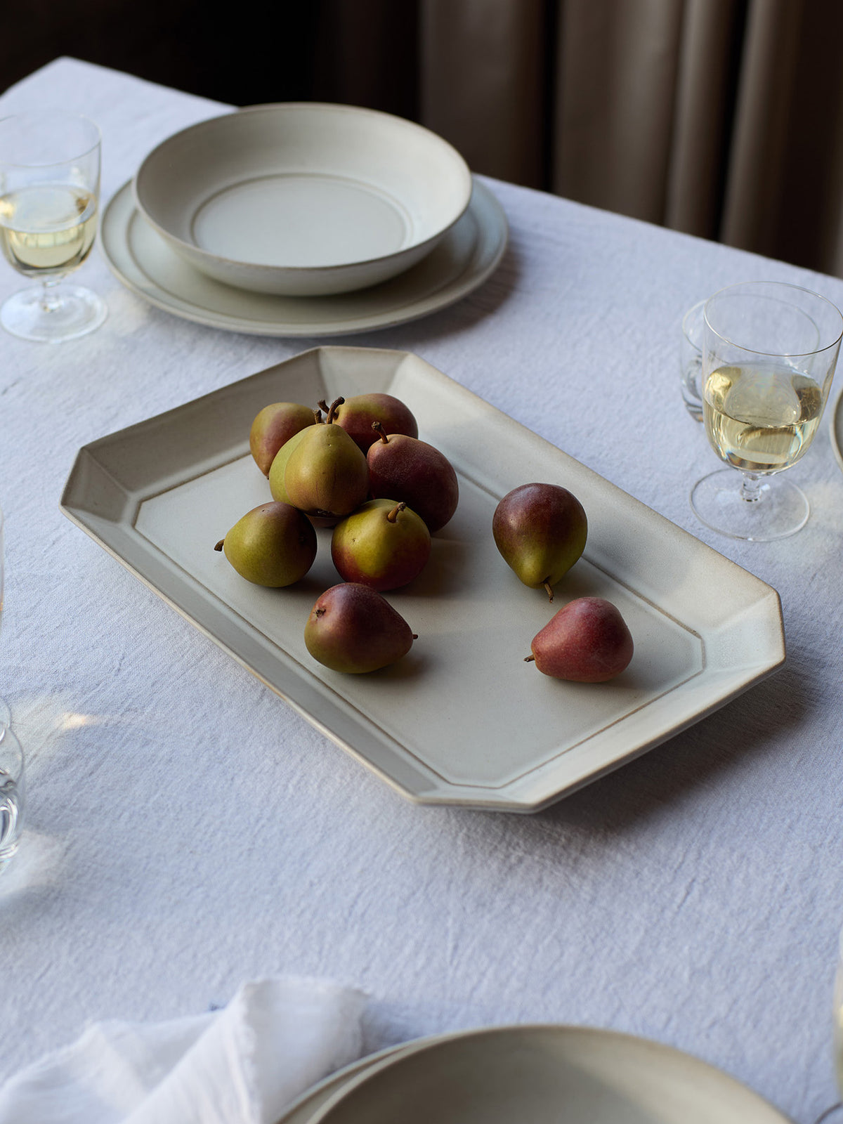 A white rectangular ceramic serving platter with a faceted rim, styled on a white tablecloth with pears on top, alongside matching plates and wine glasses.