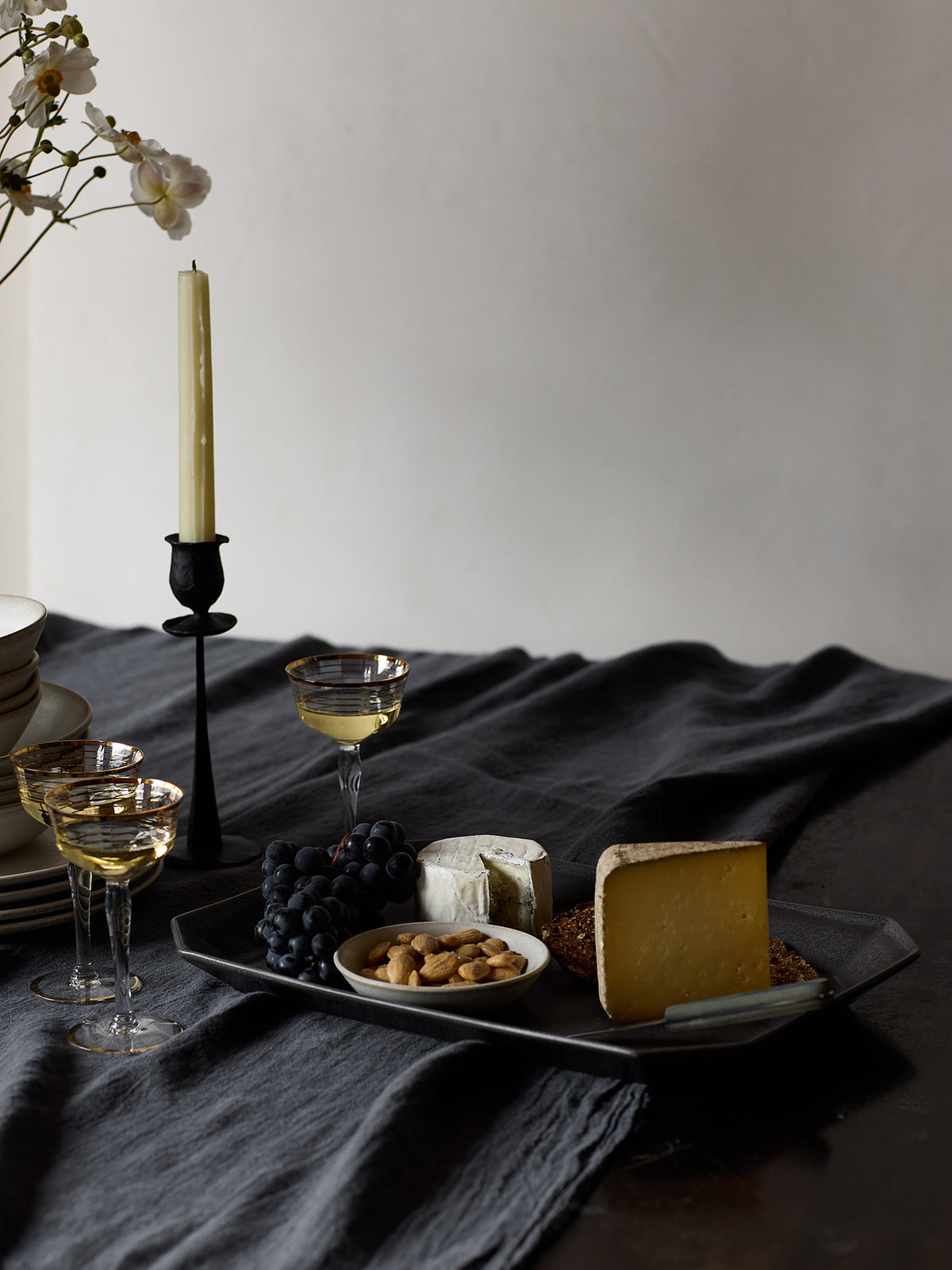 A snack spread styled on a dark grey serving platter on a dark tablecloth, featuring cheese wedges, grapes, nuts, and glasses of white wine, with a black candlestick holder and flowers in the background.