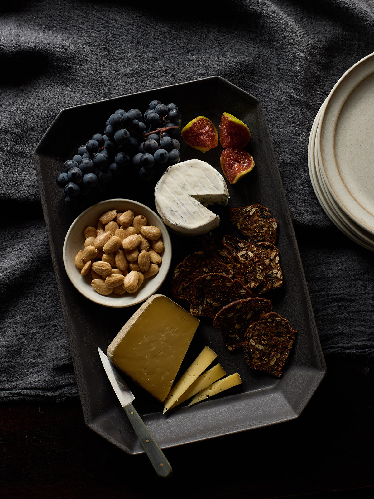 A dark gray rectangular ceramic serving platter styled with an assortment of food, including a wedge of brie cheese, a slice of cheddar, crackers, blueberries, figs, and almonds, set on a dark textured cloth.