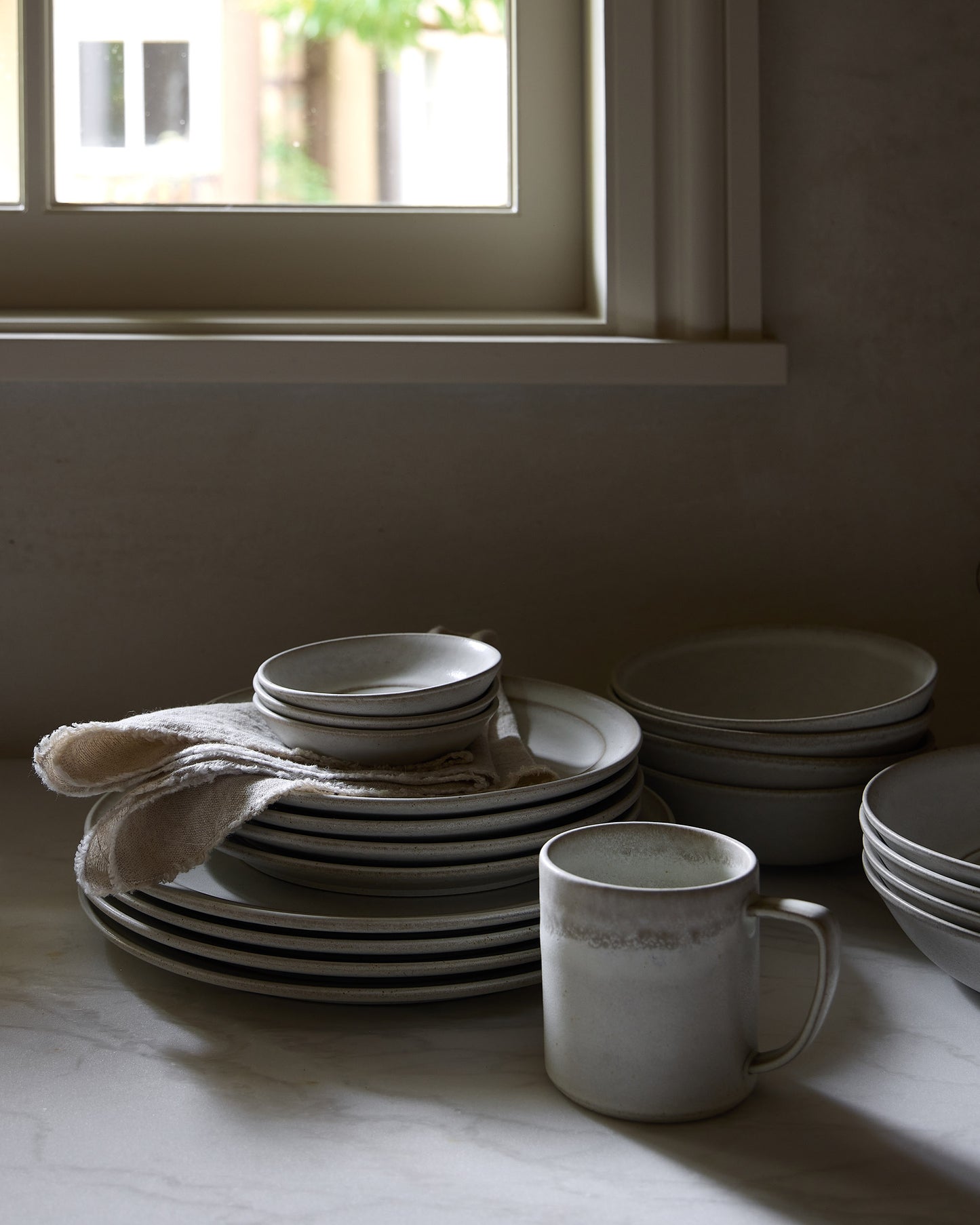 Stacks of ceramic plates and bowls on a countertop with a window in the background