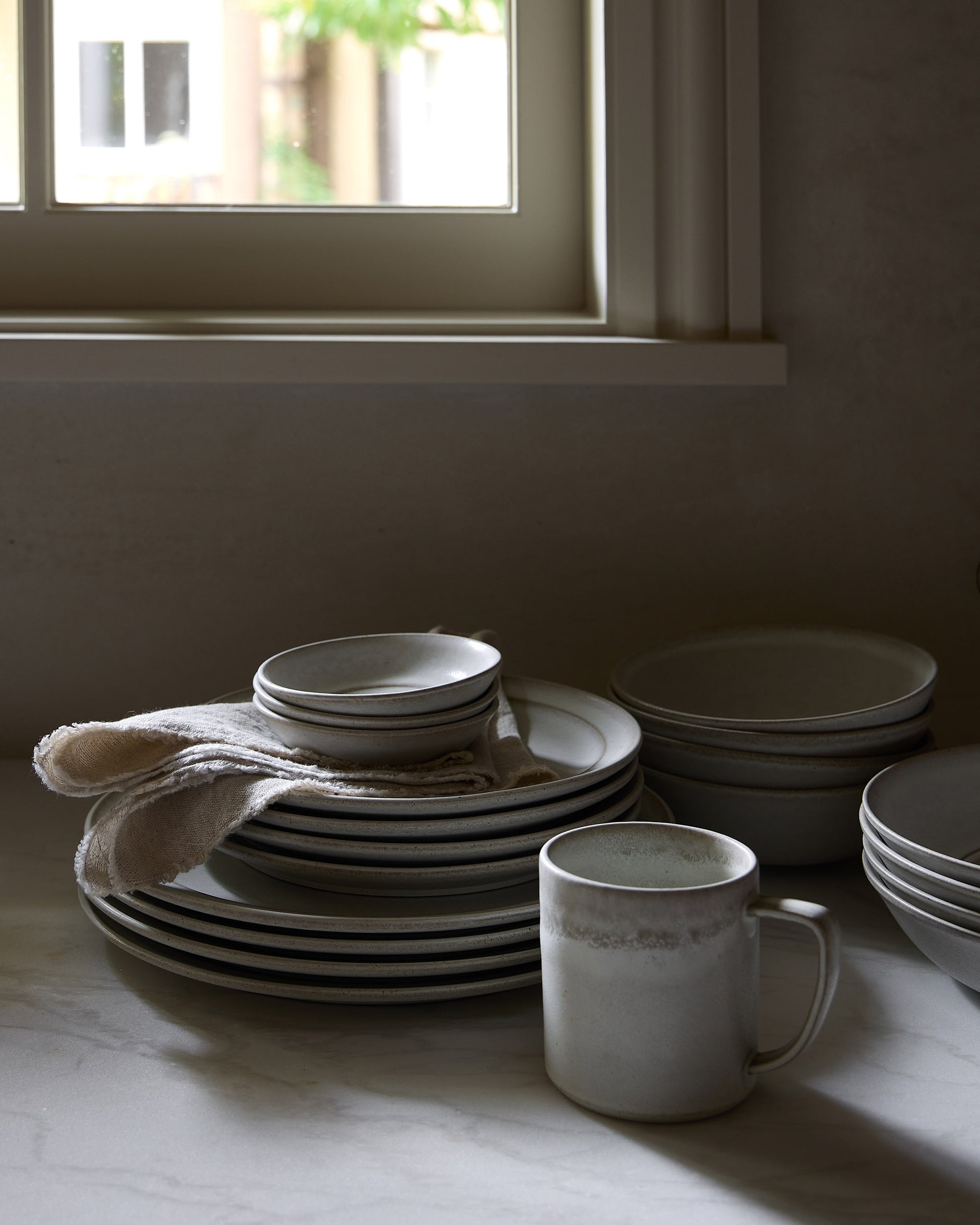 Stacks of ceramic plates and bowls on a countertop with a window in the background