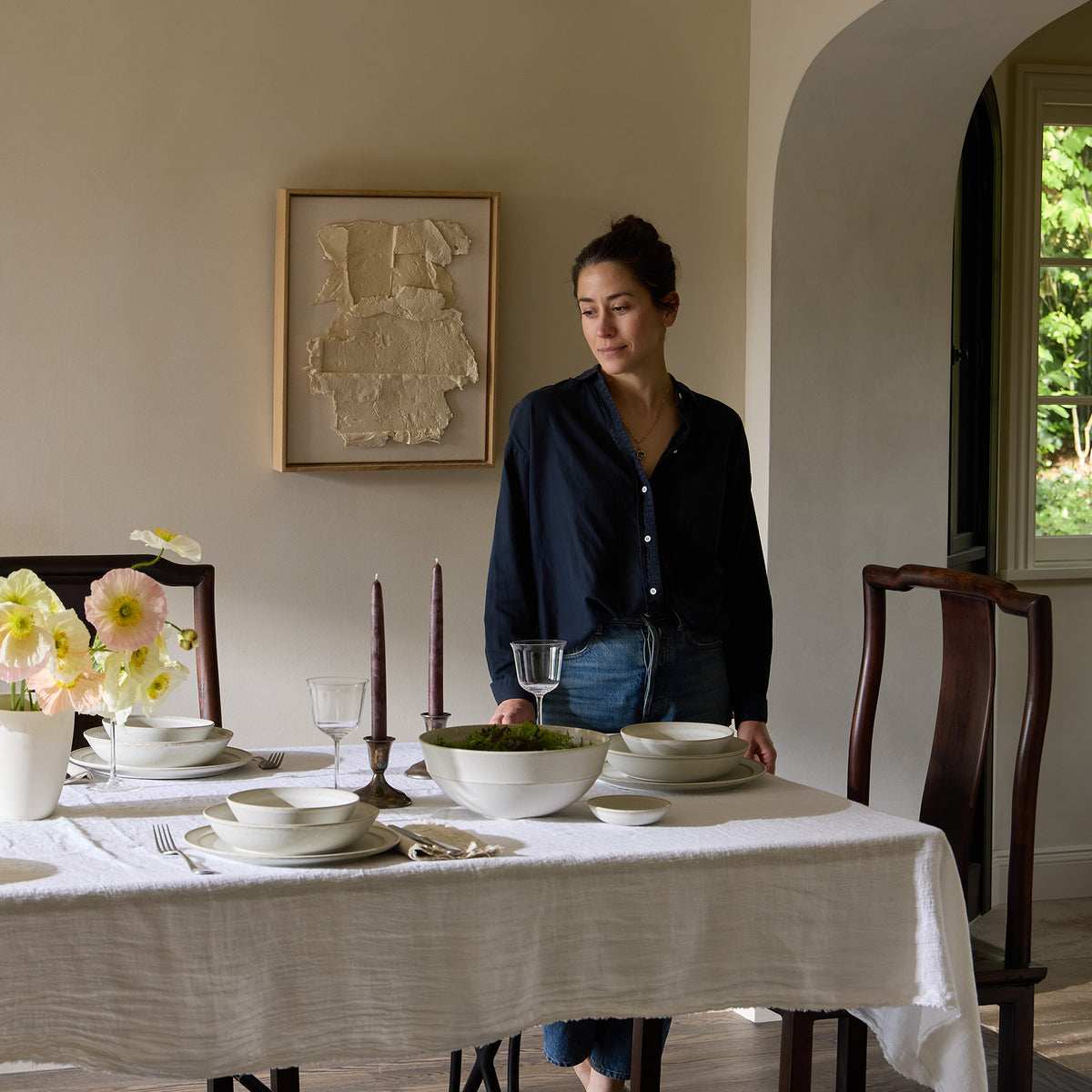 A woman stands beside a dining table set with a white tablecloth, white plates, glassware, and dark purple taper candles in a softly lit room with beige walls and framed wall art.