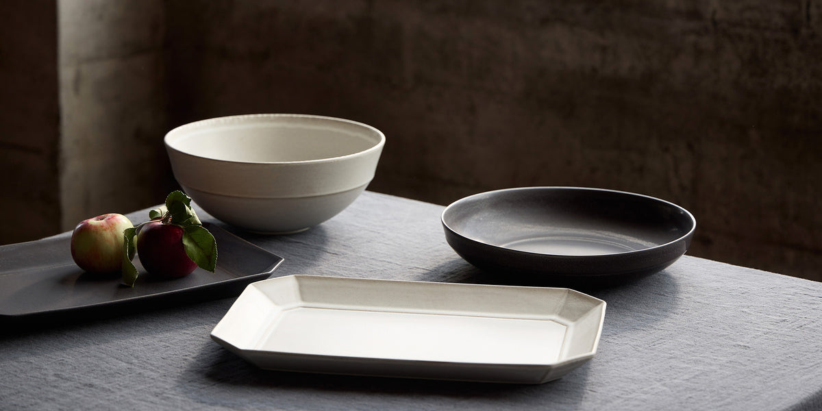 A set of minimalist ceramic tableware including a rectangular platter, a round shallow bowl, and a deep salad bowl, styled on a dark tablecloth with apples against a rustic textured background.