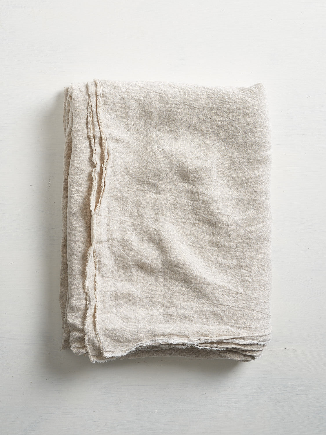 A natural linen tablecloth with a subtle jacquard grid pattern and fringed edges folded on a white surface.