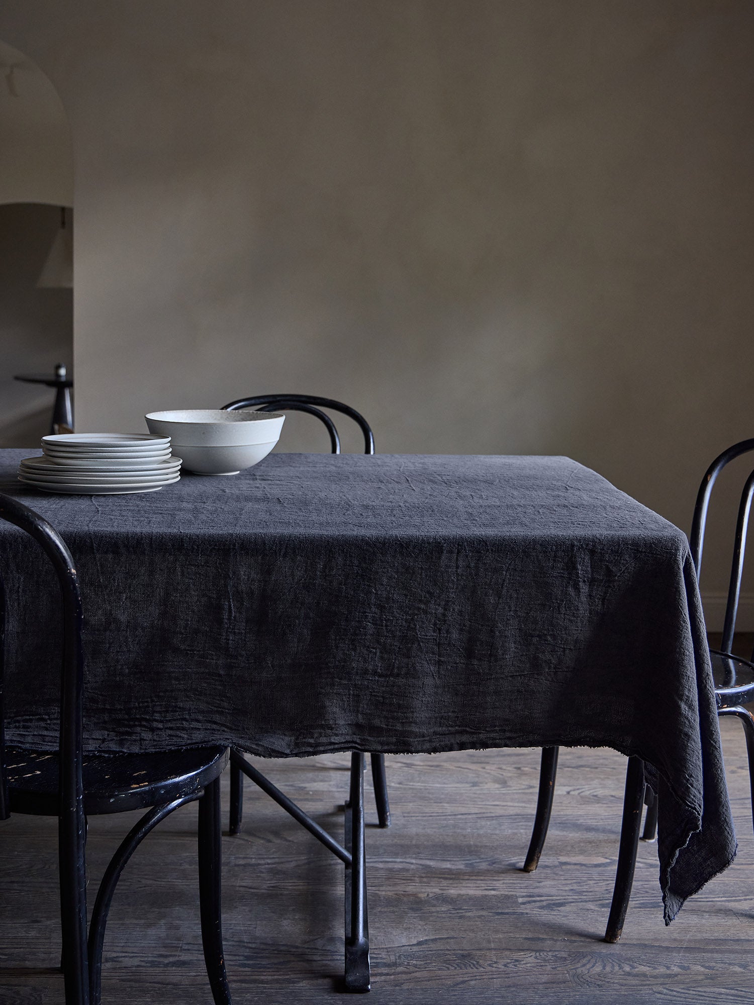 Dining table with a dark grey linen tablecloth and black chairs against a Roman clay wall.