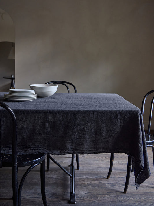 Dining table with a dark grey linen tablecloth and black chairs against a Roman clay wall.
