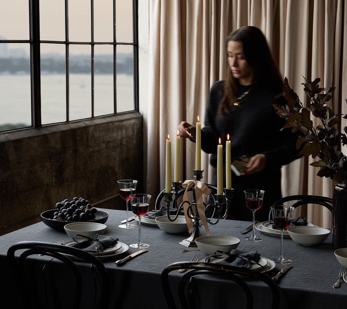 A woman in a black outfit stands beside a elegantly set dining table with lit taper candles, wine glasses, plates, and bowls in a moody room with large windows and beige curtains.